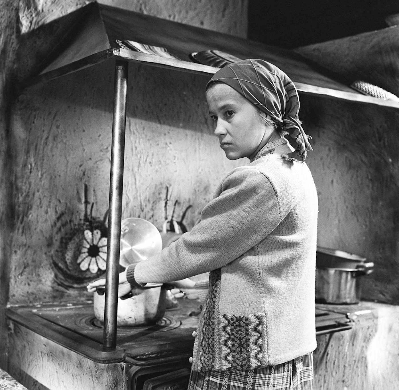 Huivipäinen nainen (näyttelijä Tarja-Tuulikki Tarsala) laittaa ruokaa puuhellalla ja katsoo sivulleen suu hieman mutrussa.