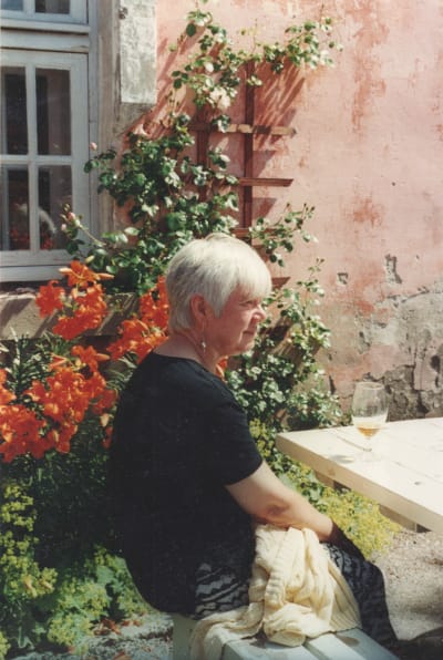 Nainen istuu puoliksi selin kameraan aurinkoisella ravintolan pihaterassilla vaaleanpunaisen seinän edessä.