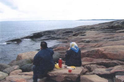 Kaksi henkilöä istuu selin kameraan rantakallioilla ja katselee avomerelle kahvia juoden ja eväitä syöden.