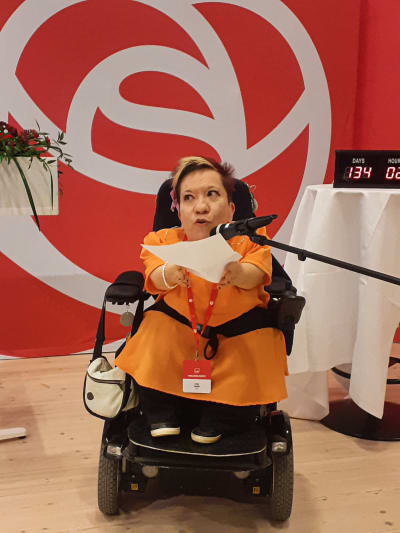 En rullstolsburen kvinna pratar i mikrofonen på en scen med SDP:s logga i bakgrunden.