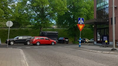 En T-korsning mellan två gator. Bilar kör och andra bilar står parkerade bakom korsningen. I bakgrunden en grön bank och gröna lövträd. Trafikmärken. 
