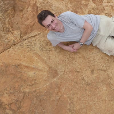 Tretåigt dinosauriefotspår i sten, bredvid ligger en glad man och poserar.