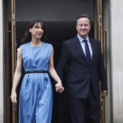 David Cameron on pukeutunut tummaan pukuun ja sinineen kravattiin. Samanthalla on yllään sininen mekko ja musta vyö.