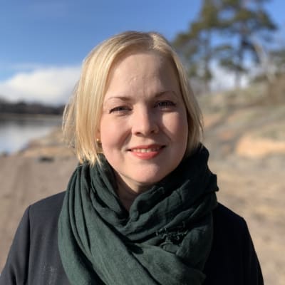 Närbild av en blond kvinna som står på en klippig strand i solskenet.