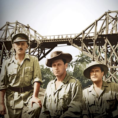 Kolme miestä sotilaspuvuissa takanaan iso puinen silta viidakossa.