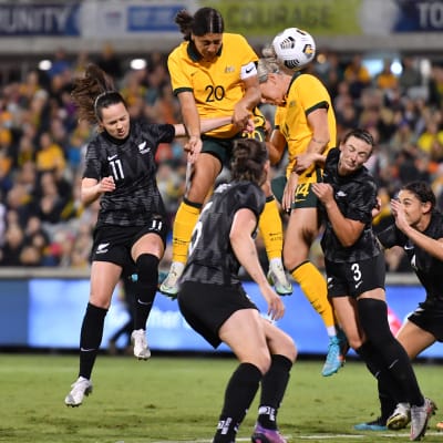 Sam Kerr når högst upp på en höjdboll. Hon är klädd i orange och omringad av svartklädda nyzeeländska spelare.