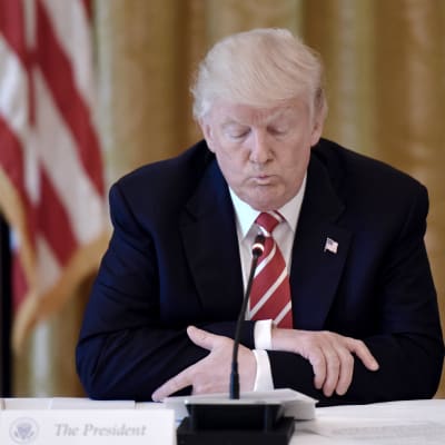 Donald Trump sitter vid ett bord med en mikrofon framför sig, med händerna knäppta och blicken vänd nedåt.
