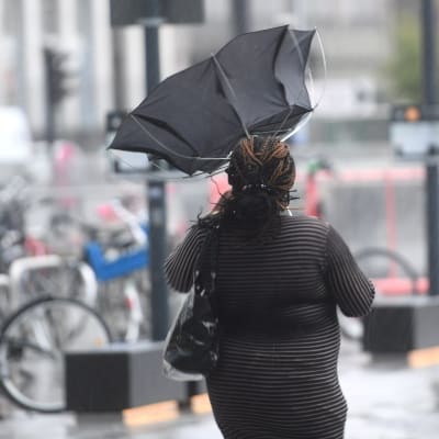 En kvinna går på gatan i grått regnväder. Hennes paraply står rakt upp efter att ha gått sönder.
