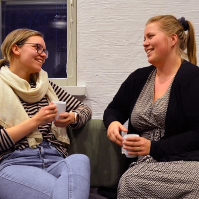 Två unga kvinnor sitter och dricker kaffe på en soffa framför en vägg.