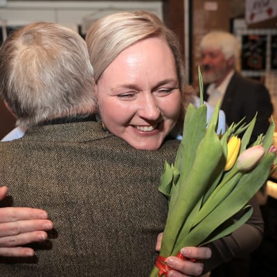 Pia Sillanpää hymyilee onnellisena tulppaanit käsissä kun häntä halaa harmaahiuksinen henkilö.