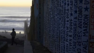 Muren som delar Mexico och USA. Ansikten och namn står inskrivna på muren. I bakgrunden syns en sandstrand och havet.