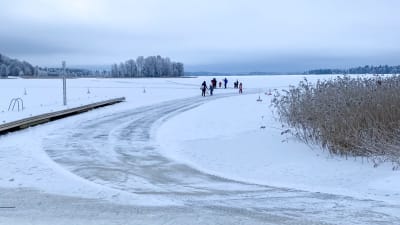 Flera personer syns ute på isen. Till vänster en brygga, till höger vass.
