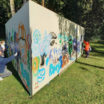 En triangelformad vägg eller ett plank som placerats ute på en gräsmatta. Barn och ungdomar målar på det.
