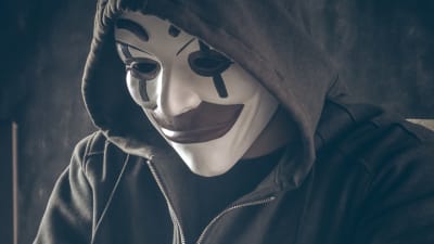 en anonym person i huva och harlekinmask sitter och skriver vid en dator.