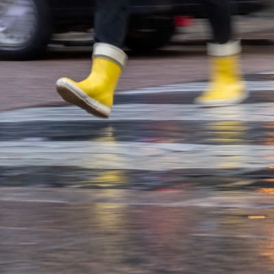 Kuvassa näkyvät sateella suojatietä ylittävät jalat, joissa on keltaiset kumisaappaat.