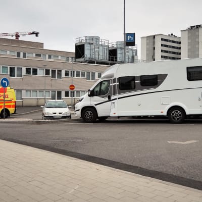 kelta-punainen ambulanssi ajaa tiilirakennuksen ja autojen pysäköuntialueen välissä Oulun yliopistollisen keskussairaalan etualalla. Pysäköintialuella valkoinen matkailuauto.