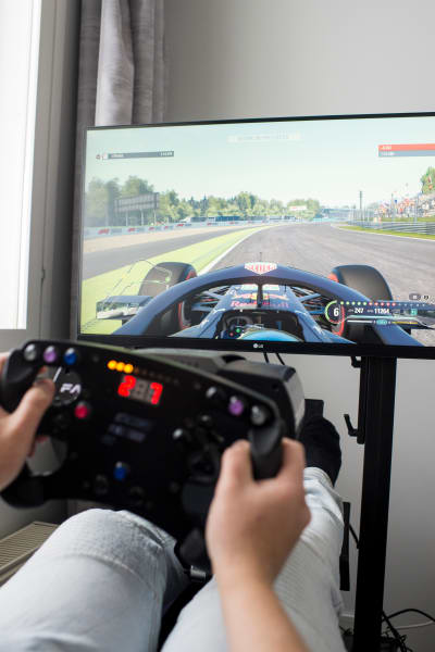 En tv-skärm som visar bilden av ett formelracingspel. I förgrunden två händer som håller i en ratt.