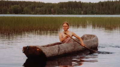 Nuori nainen meloo puunrungosta koverretulla kanootilla järvellä, taustalla sekametsäinen rinne.