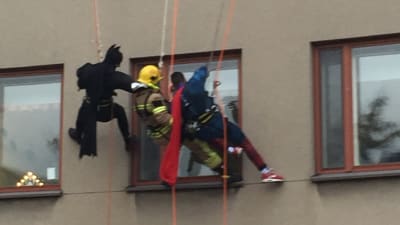 Brandmän utklädda till Batman, brandman och Stålmannen klättrar på en vägg och tittar in genom ett fönster i huset.