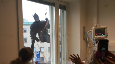Barn i ett sjukhusrum tittar ut genom fönstret och ser Batman klättra på väggen.