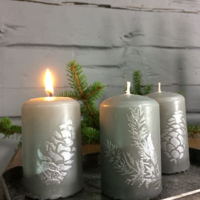 Tre ljusgråa blockljus med mönster föreställande granris och kottar i silverfärg.