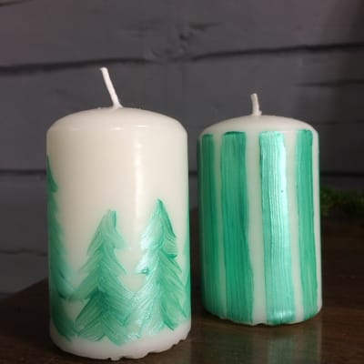 två vita blockljus det ena med mönster målat som gröna granar och det andra med målade gröna ränder.