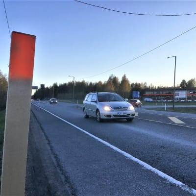En T-korsning där personbilar kör åt olika håll. Affärsbyggnader syns i bakgrunden. Asfalterad väg, skymning.