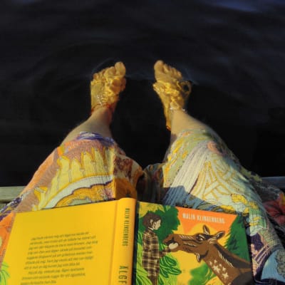 Person har fötter i vattnet och en bok i knät.