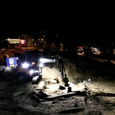 En grävmaskin med stark ledbelysning jobbar i mörkret i ett centrumområde, sand och grus syns och man kan se ljus från skyltfönster.