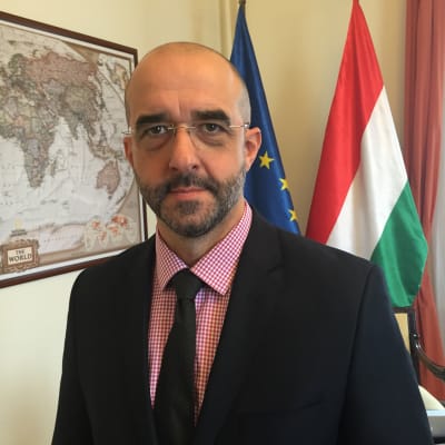 Ungerska preminärministerns talesman Zoltan Kovacs, i bakgrunden en svartvit världskart samt EU:s och Ungerns flaggor.