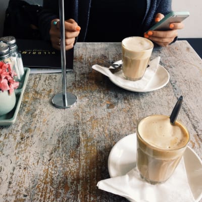 Två koppar kaffe på bordet i ett kafée och en kvinnohand som håller i en mobiltelefon.