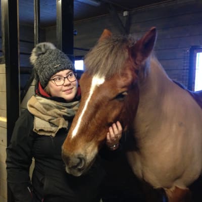 En kvinna står bredvid en häst i ett stall. Hon håller handen runt hästens mule.