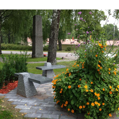 Stenbänkar och planteringar i en park. I bakgrunden en minnessten.