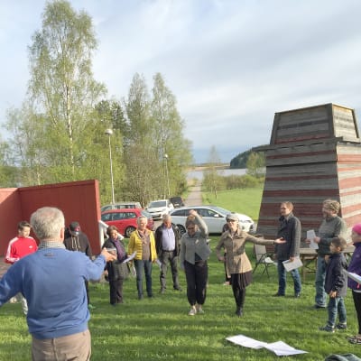 En grupp mänskor står och övar rörelser på en gräsmatta. En ledare (regissör) strö framför dem och ger instruktioner.