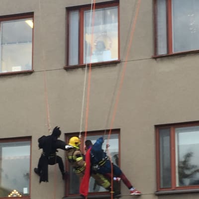 Brandmän utklädda till Batman, brandman och Stålmannen klättrar på en vägg och tittar in genom ett fönster i huset.