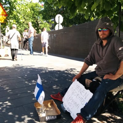 Huppariin pukeutunut suomalainen kerjäläinen istuu puun alla Helsingin Kampissa. Kerjäläisen edessä on laatikon kansi, jossa on kolikoita ja tyhjä viinapullo, jossa liehuu pieni Suomen lippu. Valkoisessa pahvissa luke: Kotimainen vaihtoehto, Kaljarahaa kiitos. Money for beers TY!
