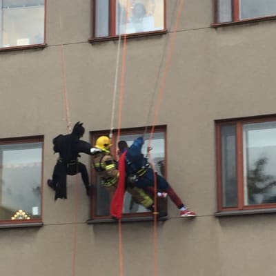 Brandmän utklädda till Batman, brandman och Stålmannen klättrar på en vägg och tittar in genom ett fönster i huset.