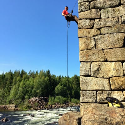 En man som klättrar upp för en stenmur med hjälp av klättringsutrustning, intill en fors.