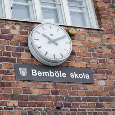 Klocka på vägg, under den skylt med texten Bemböle skola.