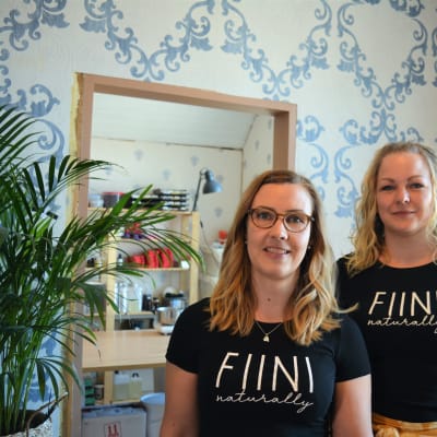 Fiinin yrittäjät Essi Rintamäki ja Heidi Sivula seisovat siniset Fiini-paidat päällä ja katsovat suoraan kameraan. Vasemmalla viherkasvi ja taustalla peili ja ornamenttitapetti.