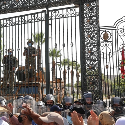Väkijoukko ja mellakkapoliiseja suljetun portin edessä, portin takana asemiehiä. 