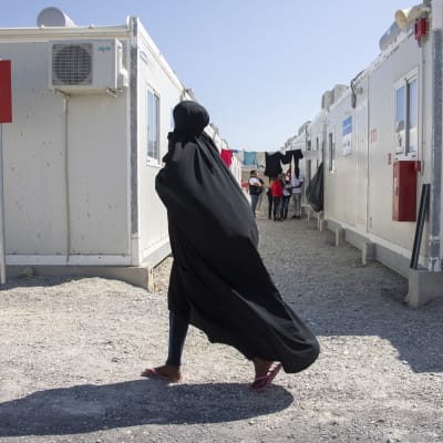 Täysin hunnutettu nainen kävelee pakolaisleirissä, takana parakkeja ja muita siirtolaisia.