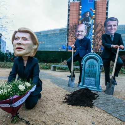 Macroniksi, Scholziksi ja von der Leyeniksi naamioituneet mielenosoittajat EU-komission edessä. Heillä on muun muassa hautakivi, jossa lukee "Lepää rauhassa EU:n Green Deal".