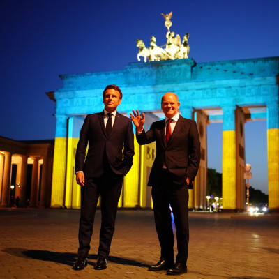 Kaksi tummiin pukeutunutta miestä ison keltasinisen portin edessä pimeässä illassa. Olaf Scholz oikealla vilkuttaa kameroille.