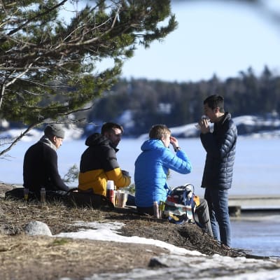 Fyra män sitter och äter matsäck på en strand. Det är vår, mend lite snö på marken. Männen är varmt klädda.