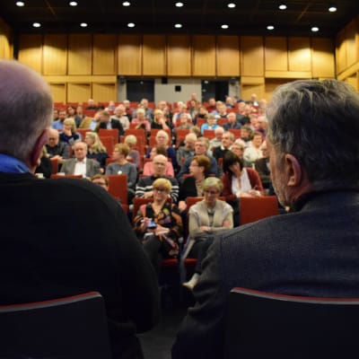 Två personer fotade bakifrån med fullsatt publik.