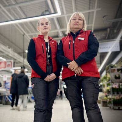 Två arbetstagare vid Citymarket står i matbutiken och tittar mot kameran. På sig har de en röd väst med affärens logo.