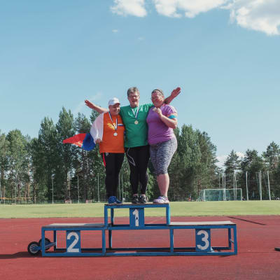 Kolme naista tuulettavat palkintokorokkeella urheilukentällä, kaulassaan mitalit.