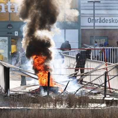 Brinnande cykelställ och annat bråte vid en gångbro till ett köpcenter. 