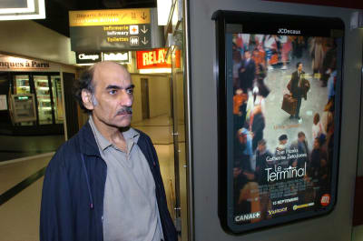 En man med buskiga ögonbryn och mustasch står bredvid en reklampelare med den franska filmaffischen för Steven Spielbergs film The Terminal.